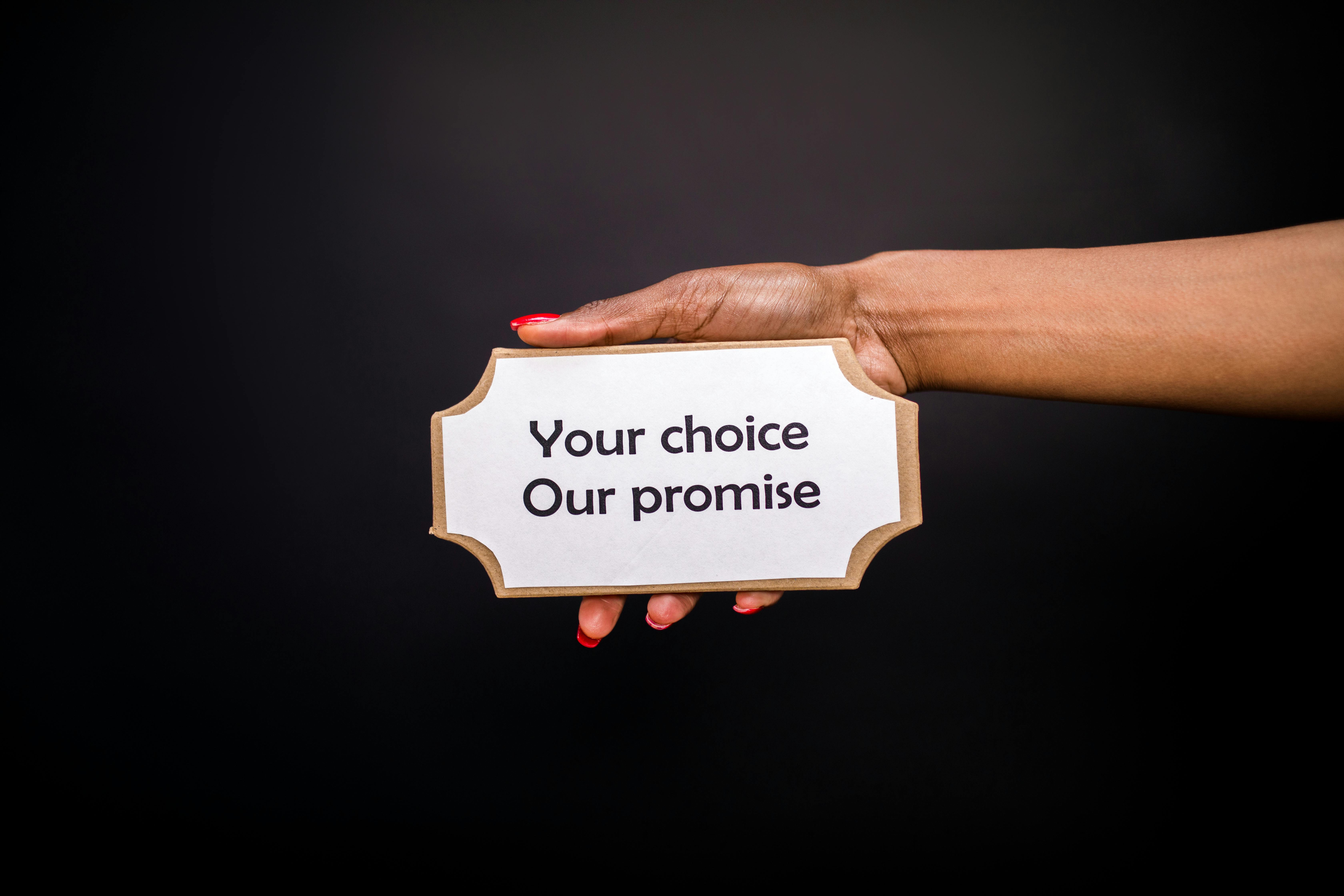 A hand holds a sign saying 'Your choice, Our promise' against a dark background.